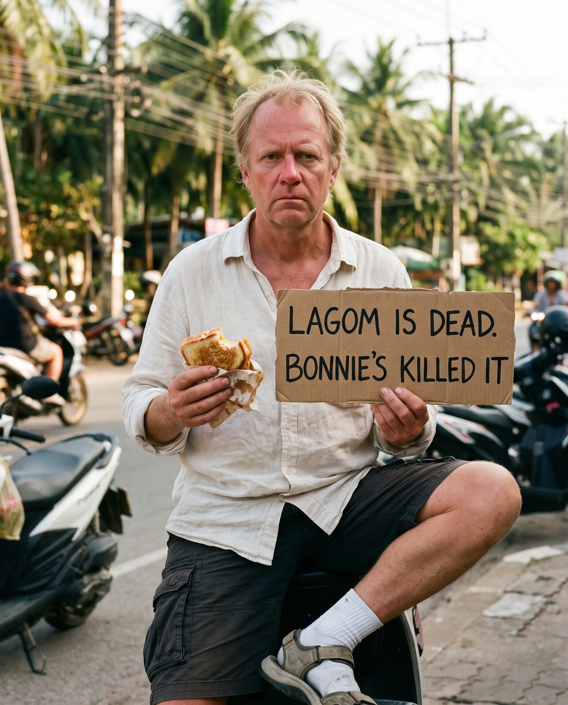 A sunburnt man in socks and sandals holding a sandwich and a sign reading Lagom is dead, Bonnie's killed it